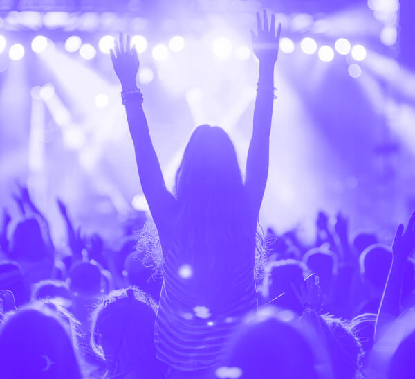 Une image de concert teintée de violet montrant une foule avec une femme au centre, les bras levés, devant une scène illuminée par des lumières vives.